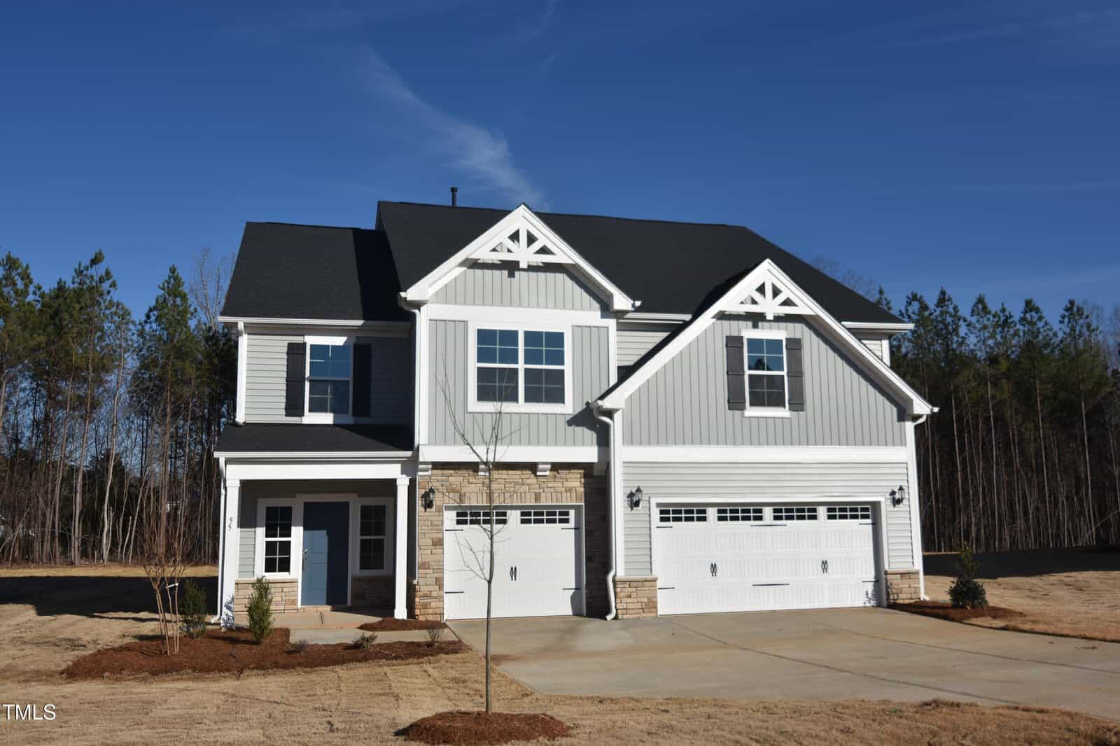 Two-story suburban house with gray siding, white trim, and stone accents. The home has a three-car garage, a covered entryway, and a large driveway. The yard is freshly landscaped with small plants and young trees.