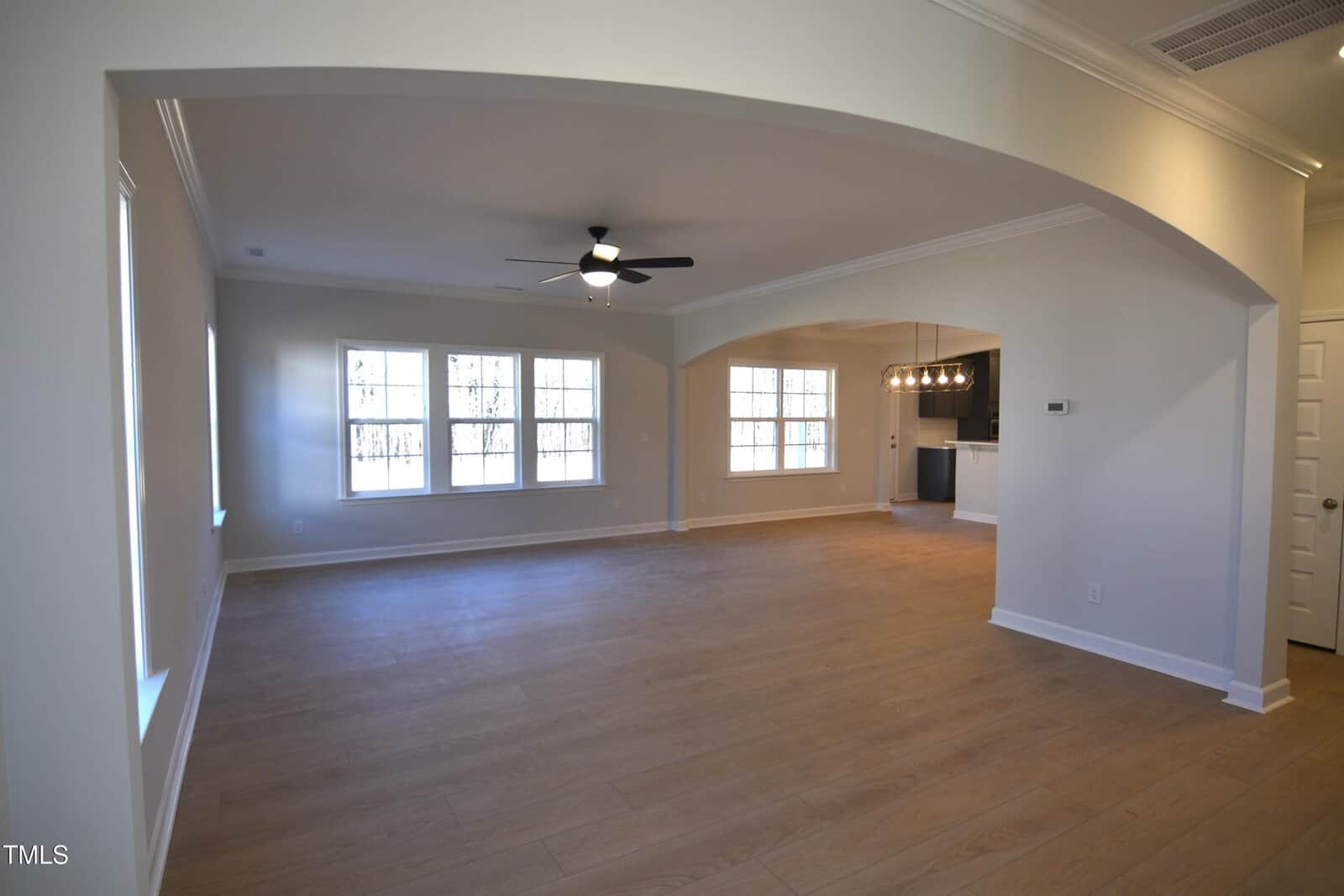 Spacious empty living room with light wood floors, large windows, ceiling fan, and arched entry. An adjacent dining area leads to a kitchen with hanging lights. Walls and ceiling are painted light neutral colors.