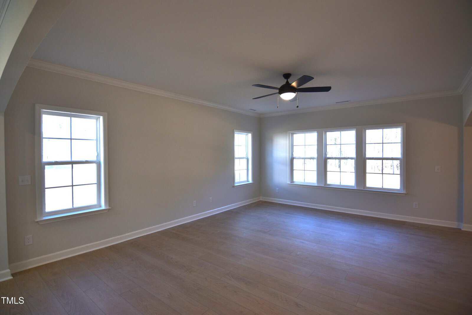 Empty living room with light wood floors, beige walls, white trim, several large windows, and a ceiling fan with a light fixture. Natural light fills the spacious area.