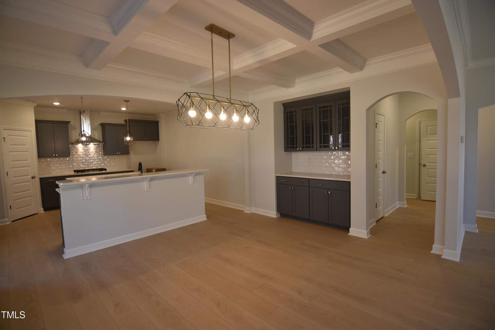 Modern open-concept kitchen and dining area with wood floors, dark cabinets, white tile backsplash, coffered ceiling, pendant lights, and arched doorway leading to a hallway with several white doors.