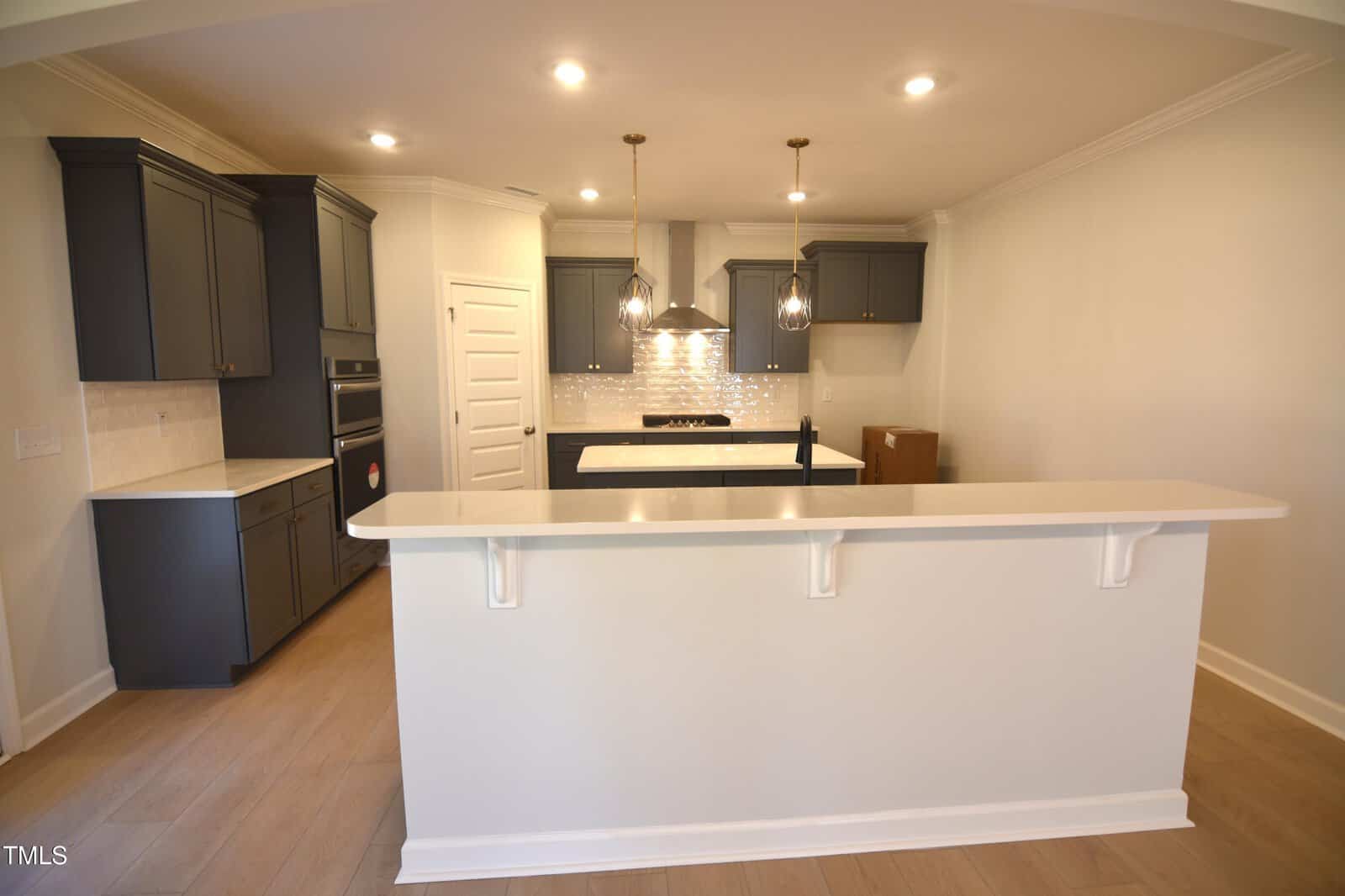 A modern kitchen with dark cabinets, light countertops, pendant lights above a white breakfast bar, stainless steel appliances, a tile backsplash, and light wood floors.