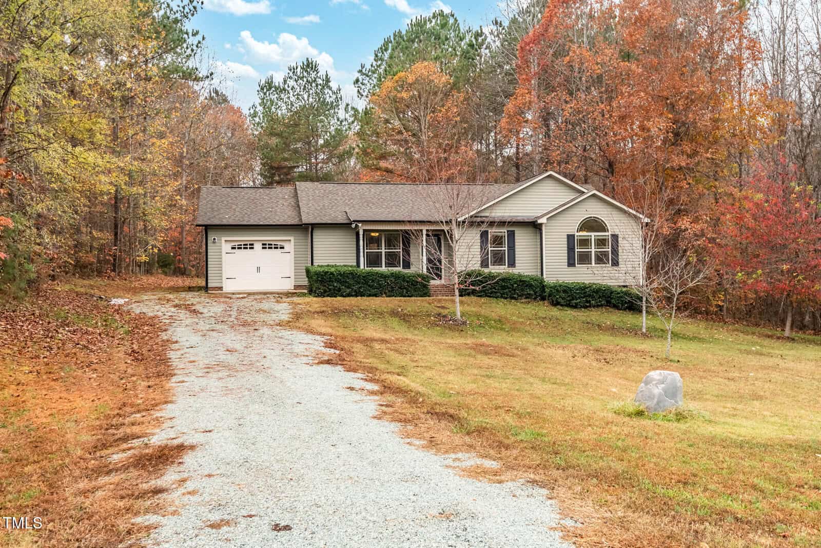 A one-story house with a beige exterior and attached garage is situated among fall foliage. The driveway is gravel, and there are trimmed bushes in front of the house. The surrounding trees display autumn colors under a partly cloudy sky.