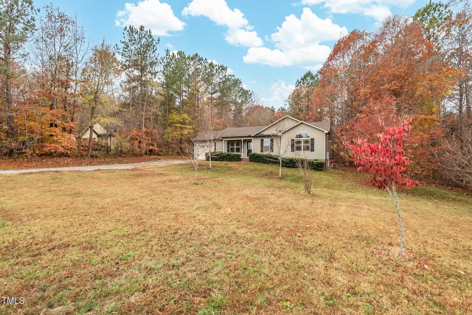 A single-story house with light-colored siding sits on a spacious grassy lawn. The surrounding trees display autumn colors, and there's a small driveway leading to the house. The sky is partly cloudy with hints of blue.