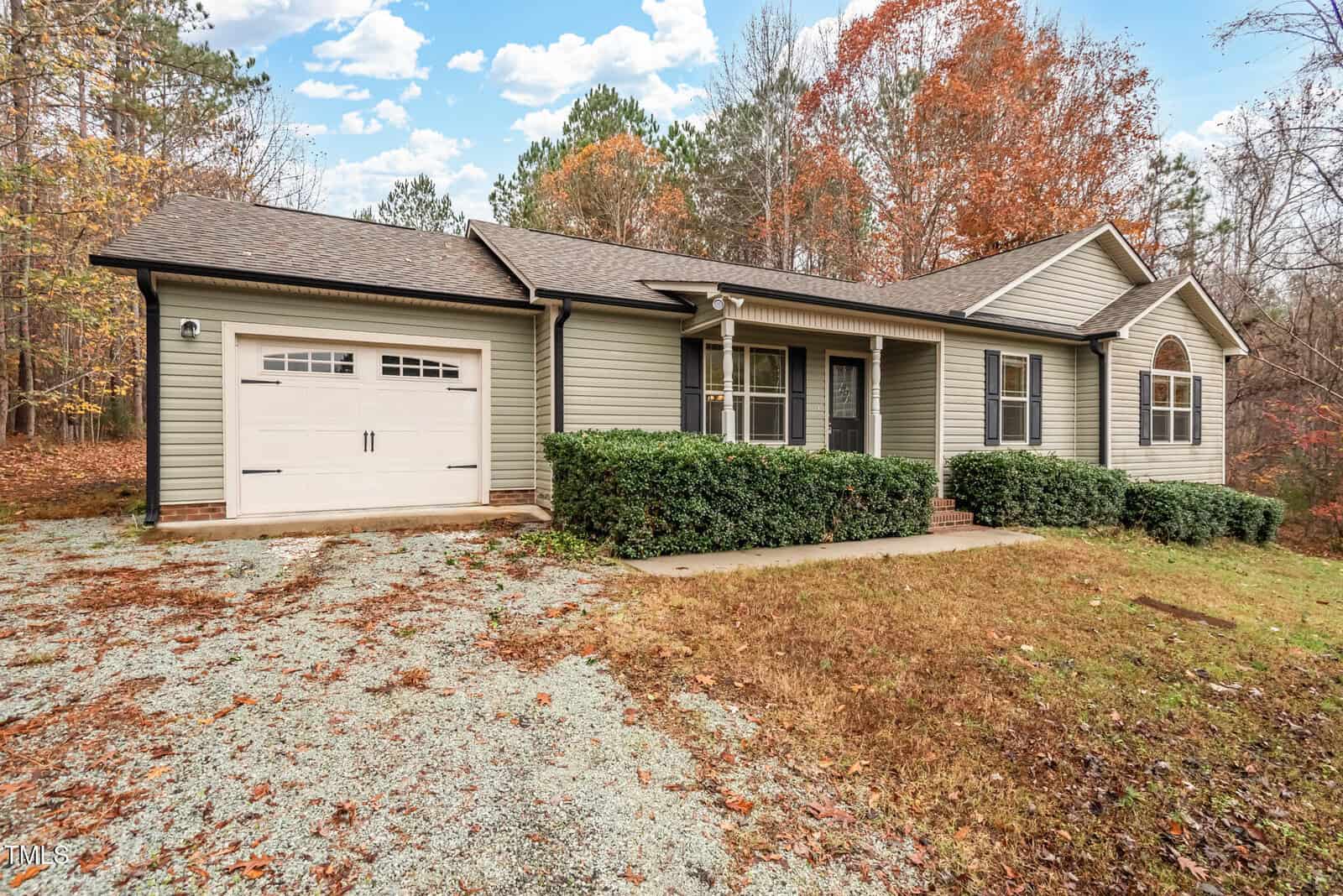 A single-story house with green siding, a one-car garage, and a front porch is surrounded by autumnal trees. The lawn is partially covered with dry leaves, and a gravel driveway leads to the garage. The sky is partly cloudy.