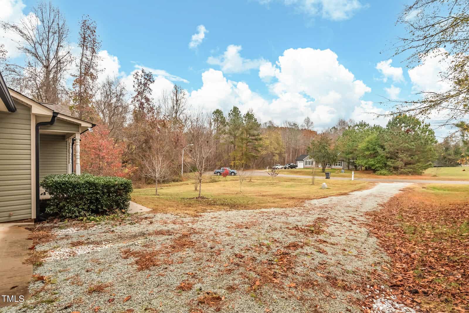 A rural outdoor scene featuring a gravel driveway leading to a house partially visible on the left. The area is surrounded by trees with autumn foliage. A few parked vehicles are seen in the distance under a partly cloudy sky.