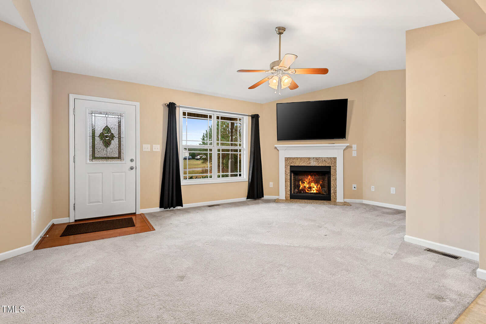 A living room with beige walls and carpet, featuring a ceiling fan and a window with black curtains. A fireplace under a wall-mounted TV is on the right. A white door with a decorative glass panel is on the left. The room is well-lit and empty.