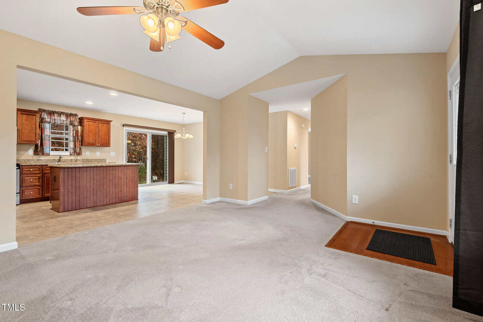 A spacious, carpeted living area with a ceiling fan leads to an open kitchen featuring wooden cabinets and tile flooring. Sliding glass doors open to the outside, and a hallway is visible to the right. Walls are painted in a neutral beige tone.