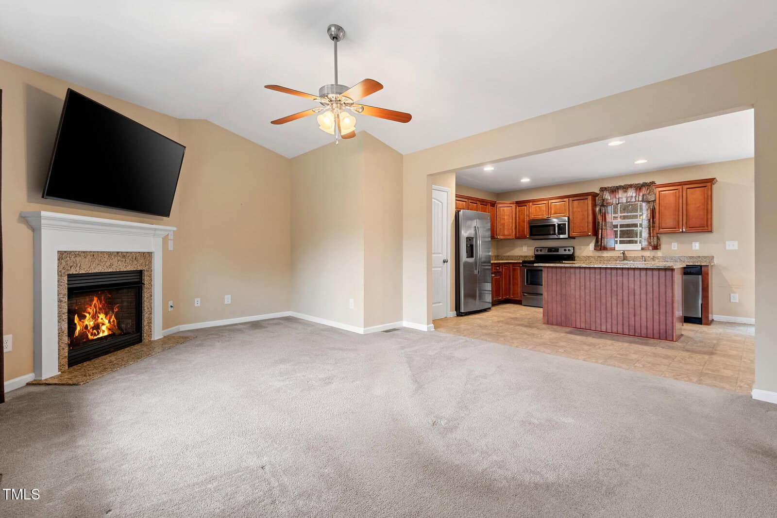 Living room with beige carpet, a ceiling fan, and a wall-mounted TV above a fireplace. Adjacent is a kitchen with brown cabinetry, stainless steel appliances, and a central island. The walls are a soft beige color.