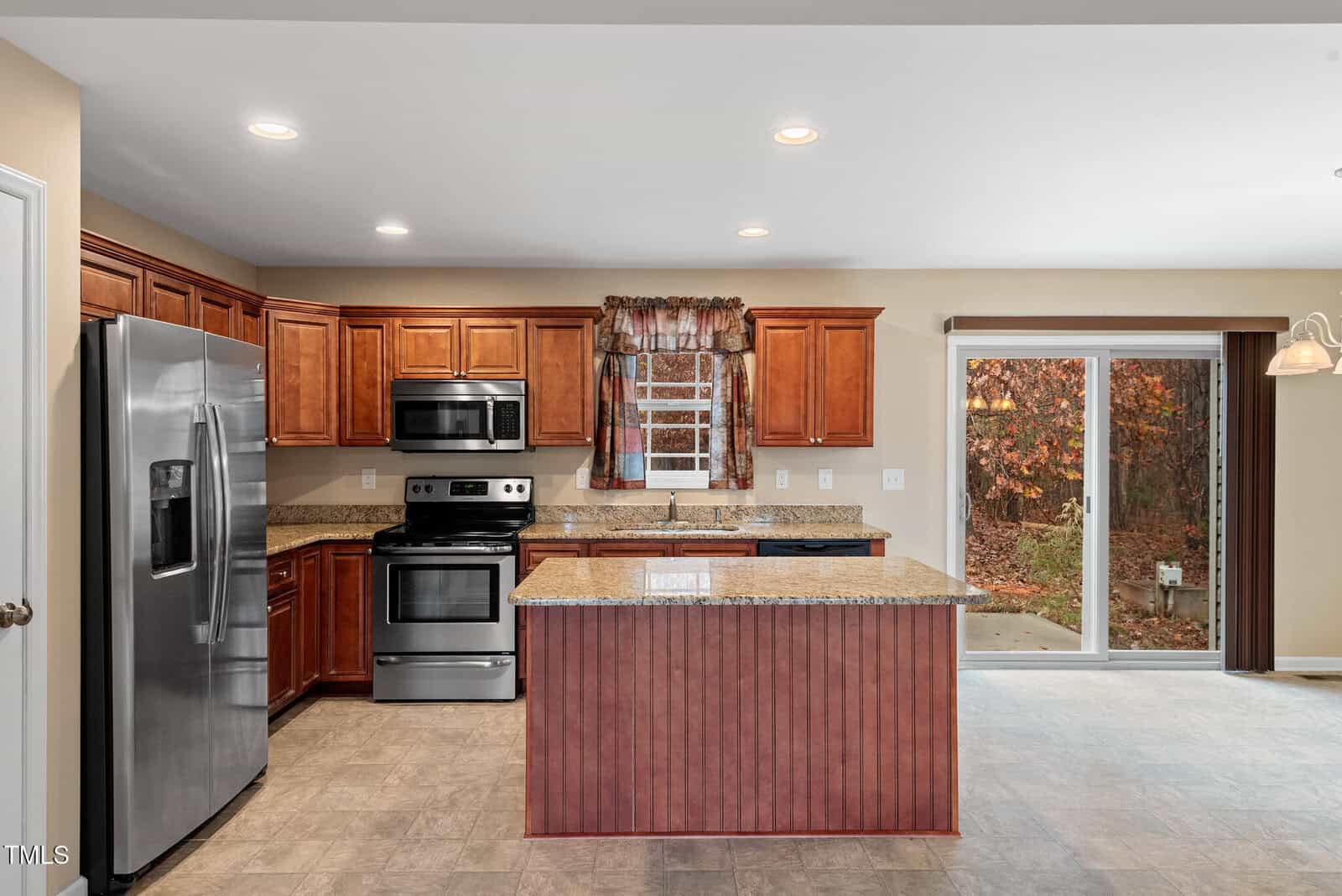 A modern kitchen with stainless steel appliances, wooden cabinets, and a central island with a granite countertop. There is a large fridge, an oven with a stove, a microwave, and a window with a floral valance. A sliding glass door opens to the outside.