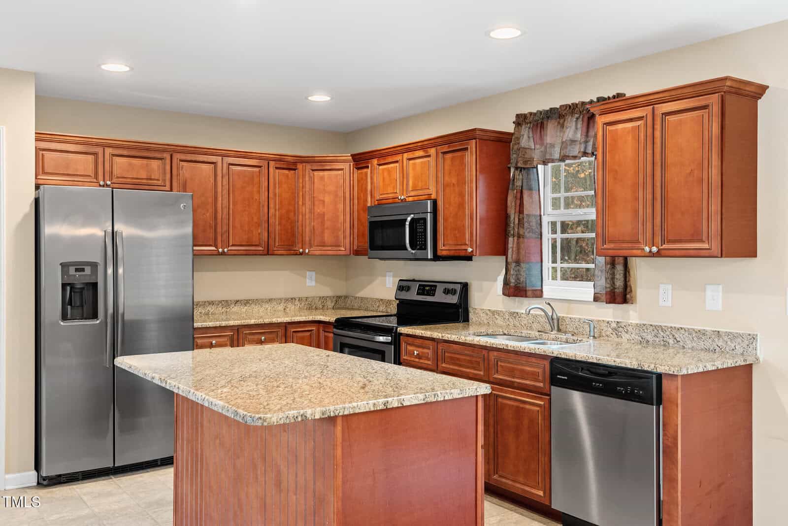 A modern kitchen features wooden cabinets, a marble island countertop, and stainless steel appliances, including a refrigerator, stove, microwave, and dishwasher. A window with patterned curtains is above the sink, and recessed lighting brightens the space.