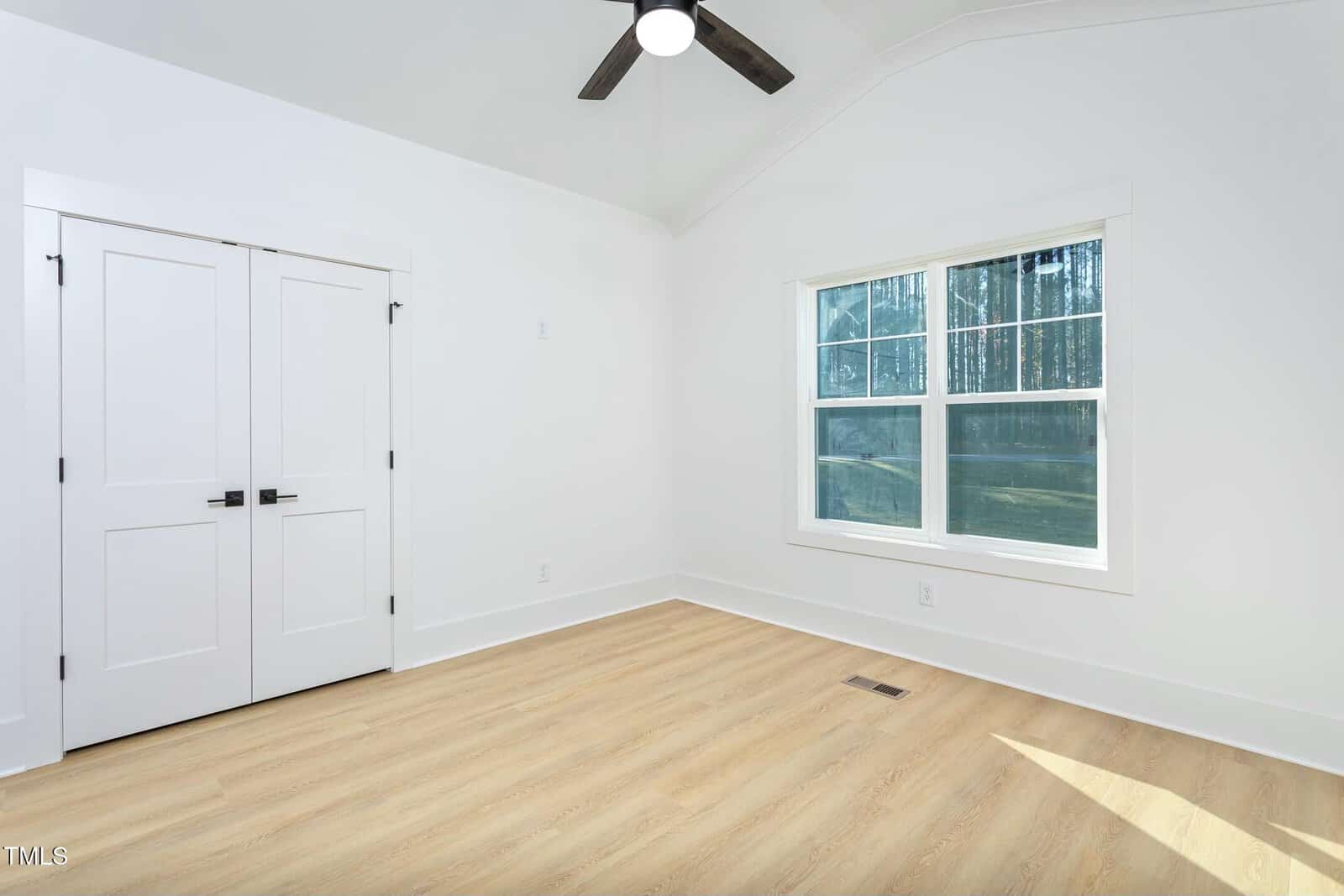 A bright and empty room features light wooden floors, white walls, and a double-door closet. A large window on the right lets in natural light. A ceiling fan is centered on the vaulted ceiling.