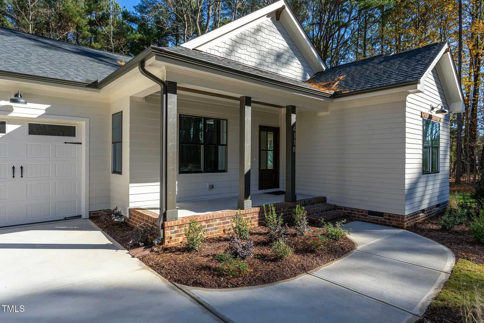 Single-story white house with dark trim sits on a concrete driveway. The front porch is supported by four columns and surrounded by small shrubs. Trees are visible in the background.