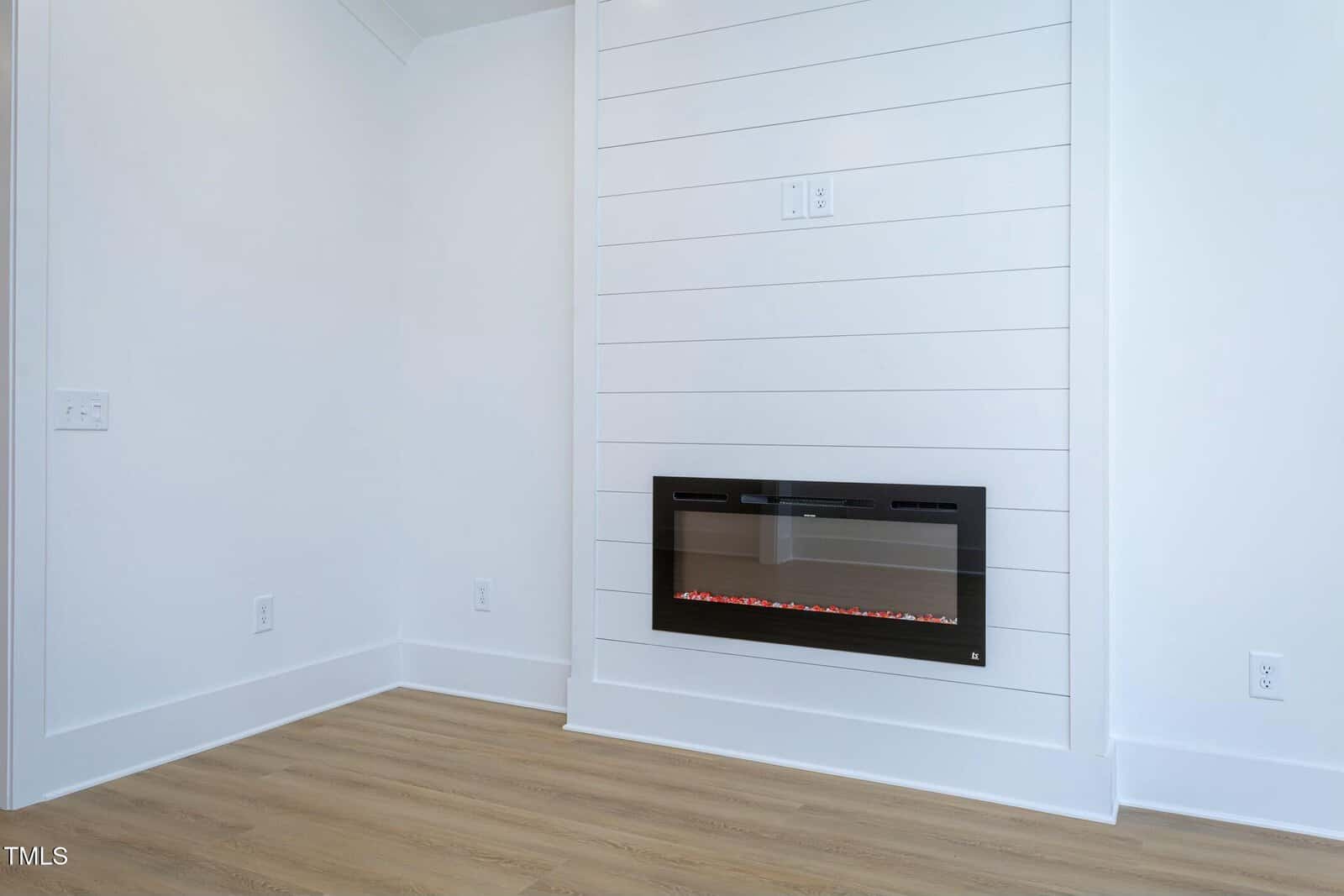 A modern, minimalist living space featuring a wall-mounted electric fireplace with a glass front. The room has light wooden flooring and white shiplap walls, creating a clean and contemporary look. Electrical outlets are visible on the walls.