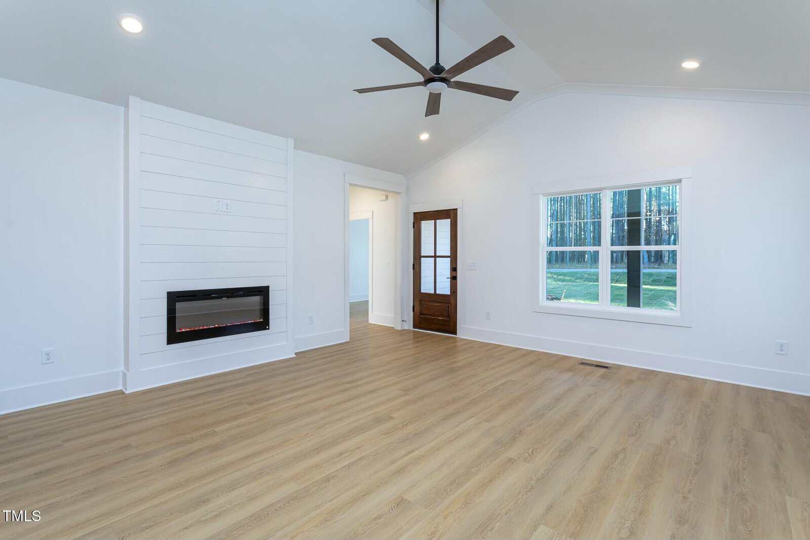 A modern, empty living room with light wood flooring and white walls. It features a wall-mounted electric fireplace, a ceiling fan, recessed lighting, and a large window. A wooden door leads to another room. Natural light enters through the window.