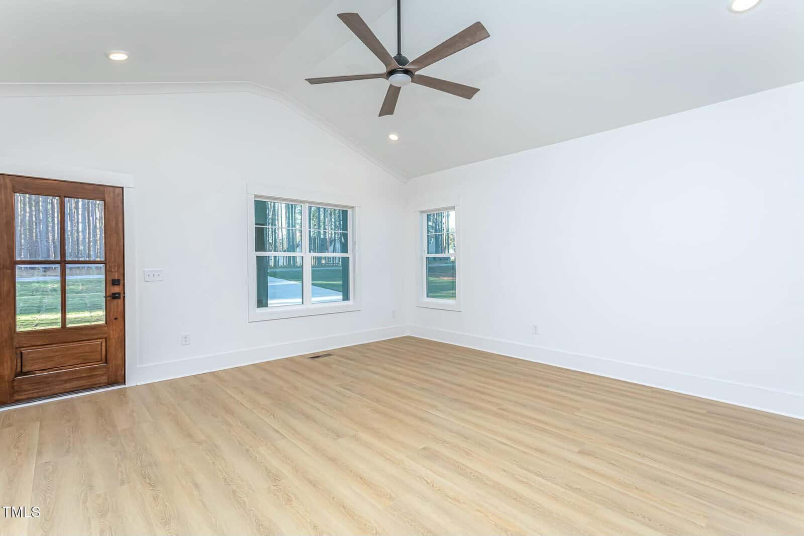A bright, empty room featuring light wood flooring, white walls, and a high ceiling with a large fan. There are two windows allowing natural light and a wooden door with glass panels on the left side.