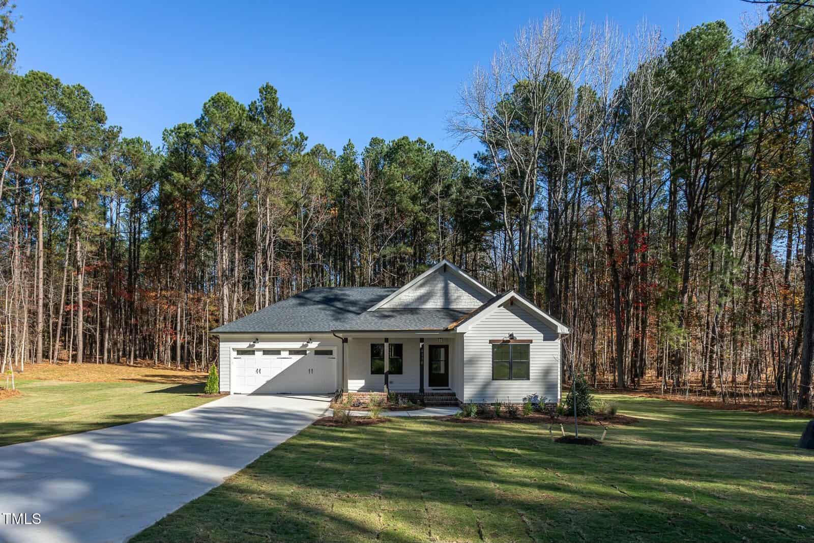 A single-story white house with a gable roof and attached garage sits in a large yard. A driveway leads to the garage. The property is surrounded by tall trees and has a clear blue sky above.