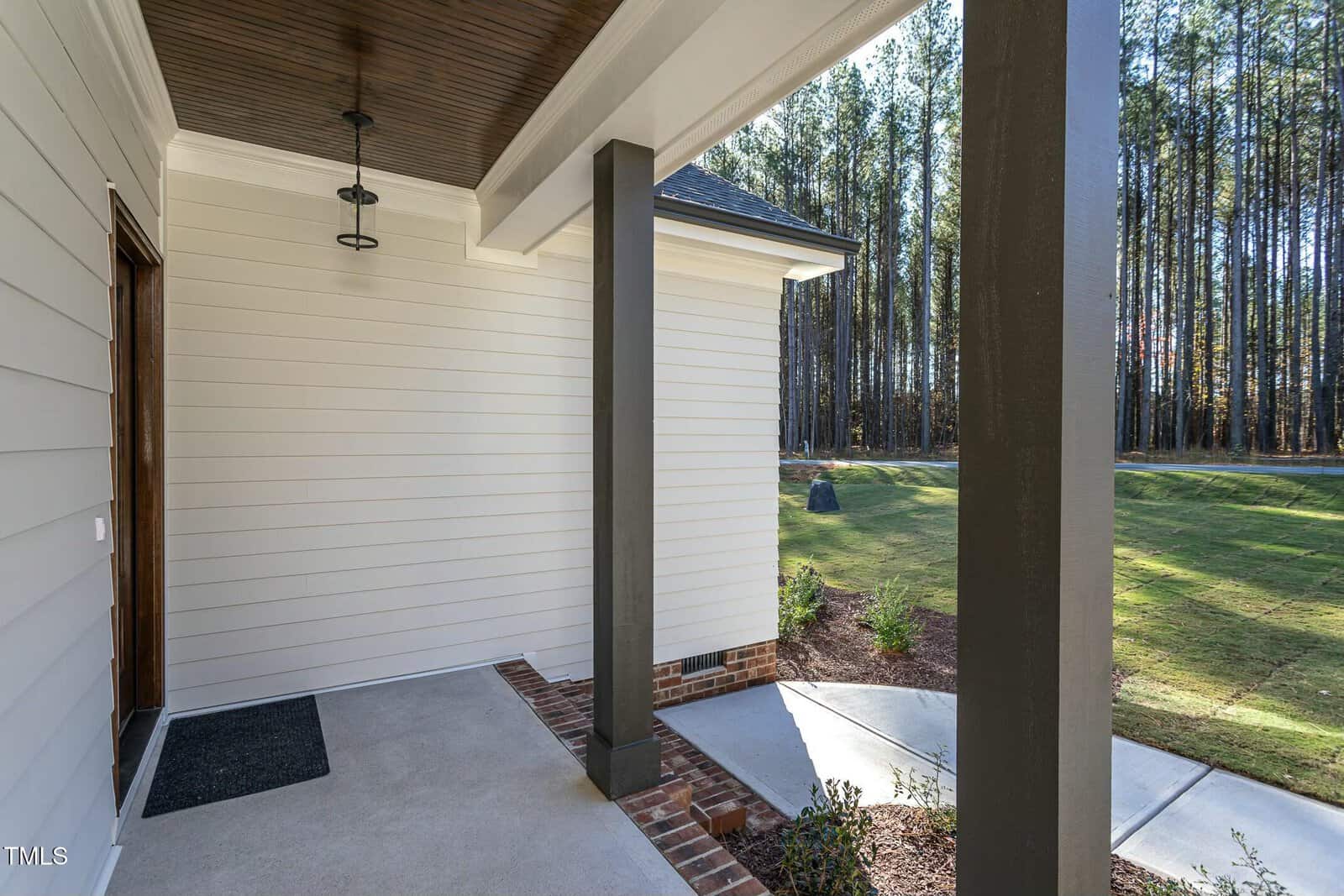 A view of a house entrance with a covered porch featuring two columns. The white exterior contrasts with a wooden ceiling and brick accents. A yard with grass and trees is visible in the background.