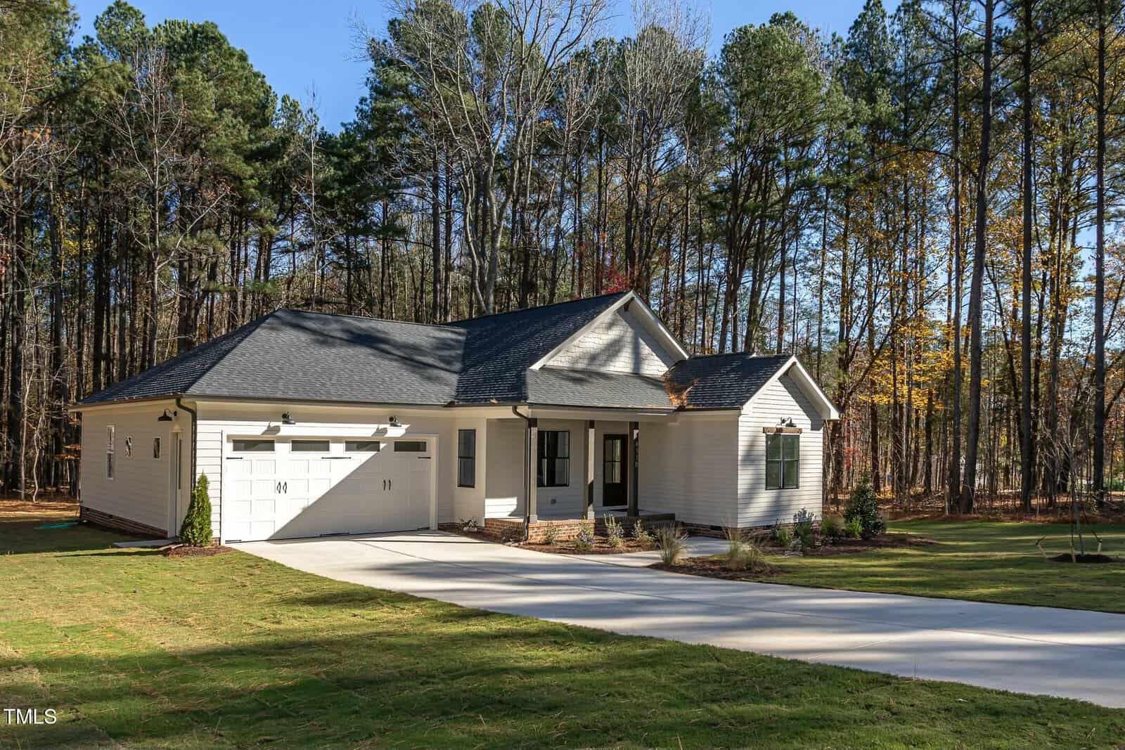 A single-story house with a light exterior sits amidst a wooded area. It features a two-car garage and a neatly maintained lawn with a concrete driveway. Tall trees provide a backdrop, and autumn leaves are scattered on the ground.