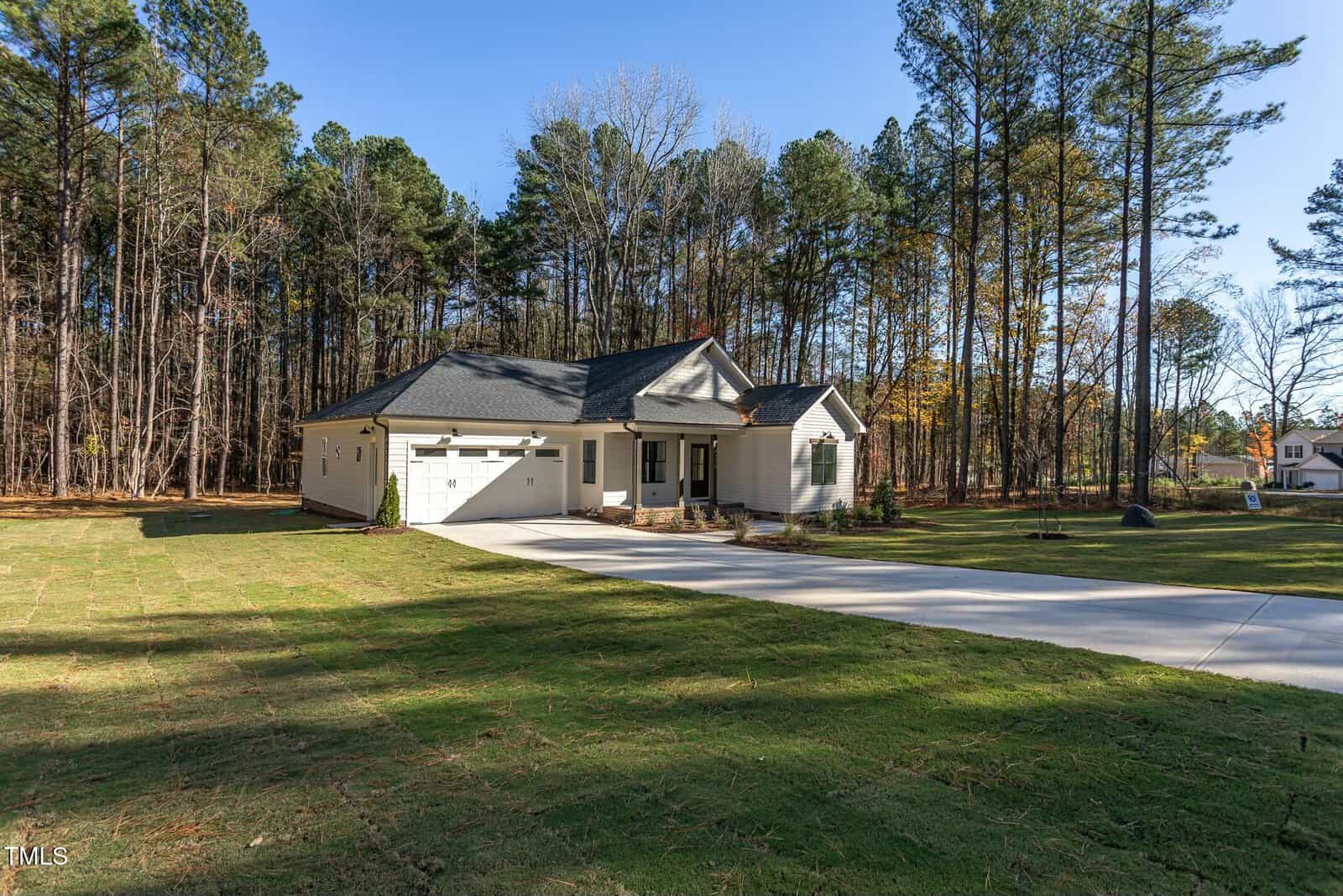 A single-story house with white siding and dark roofing is surrounded by a grassy lawn and tall trees. A long driveway leads to the garage. The scene is set on a clear day.