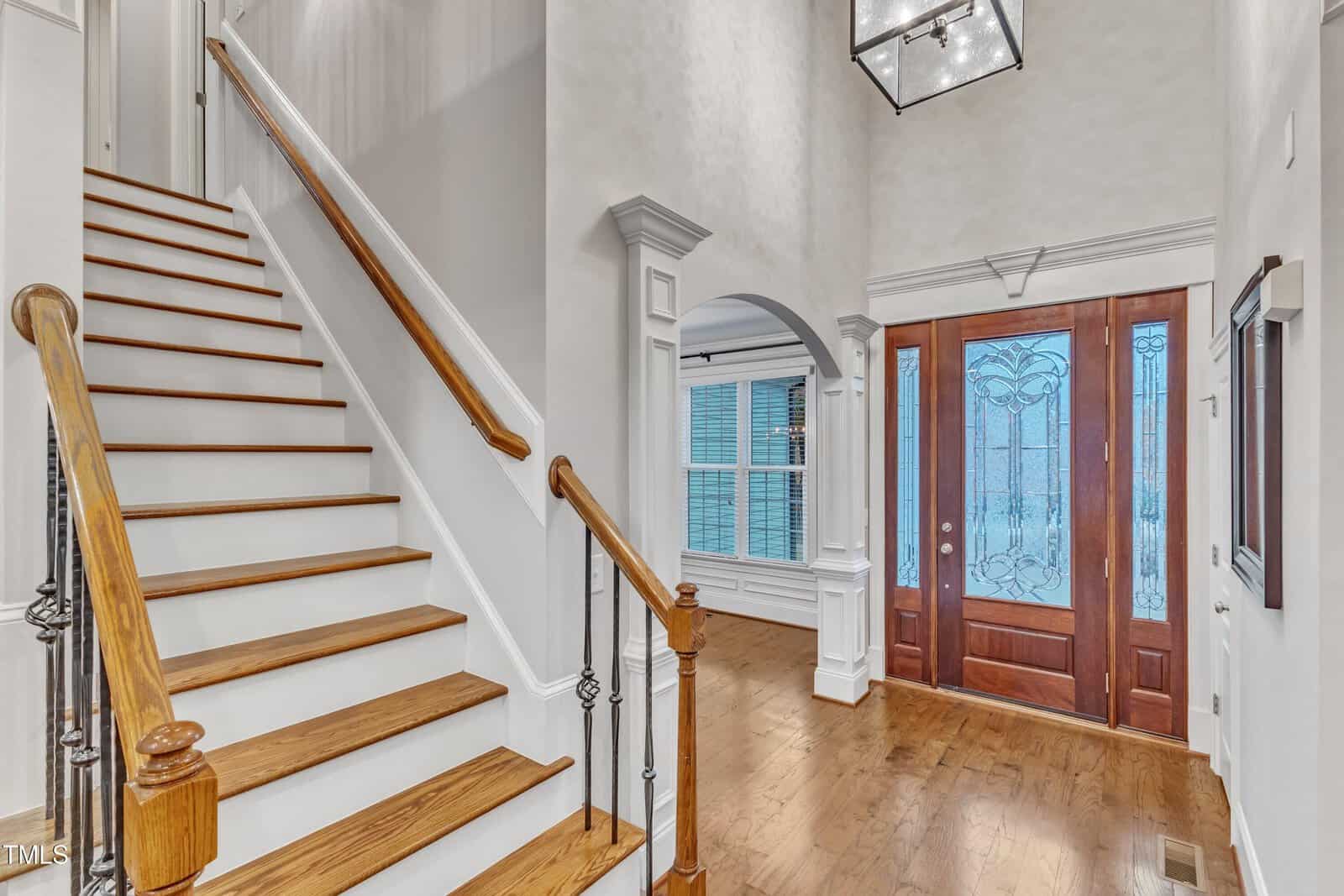 Bright entryway with wooden stairs and railing to the left, hardwood floors, an arched opening to another room, and a double wooden front door with decorative glass panels and a large light fixture above.