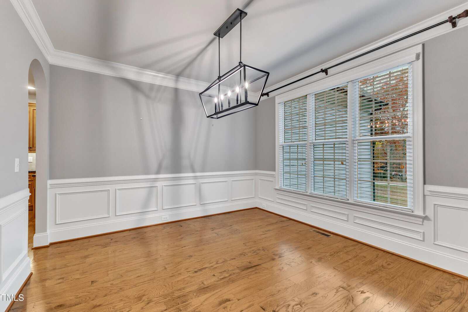 Empty dining room with light hardwood floors, gray walls with white wainscoting, large window with blinds, and a modern rectangular black chandelier hanging from the ceiling.