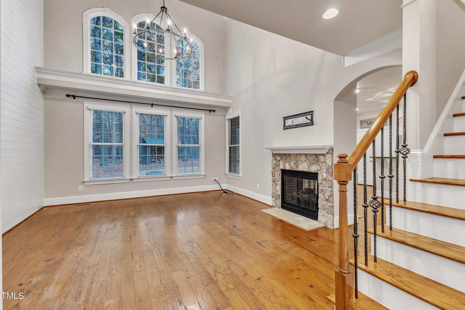 Bright living room with hardwood floors, a stone fireplace, large windows with white trim, and a high ceiling. Wooden staircase with black iron balusters is visible on the right. Room is empty and well-lit.
