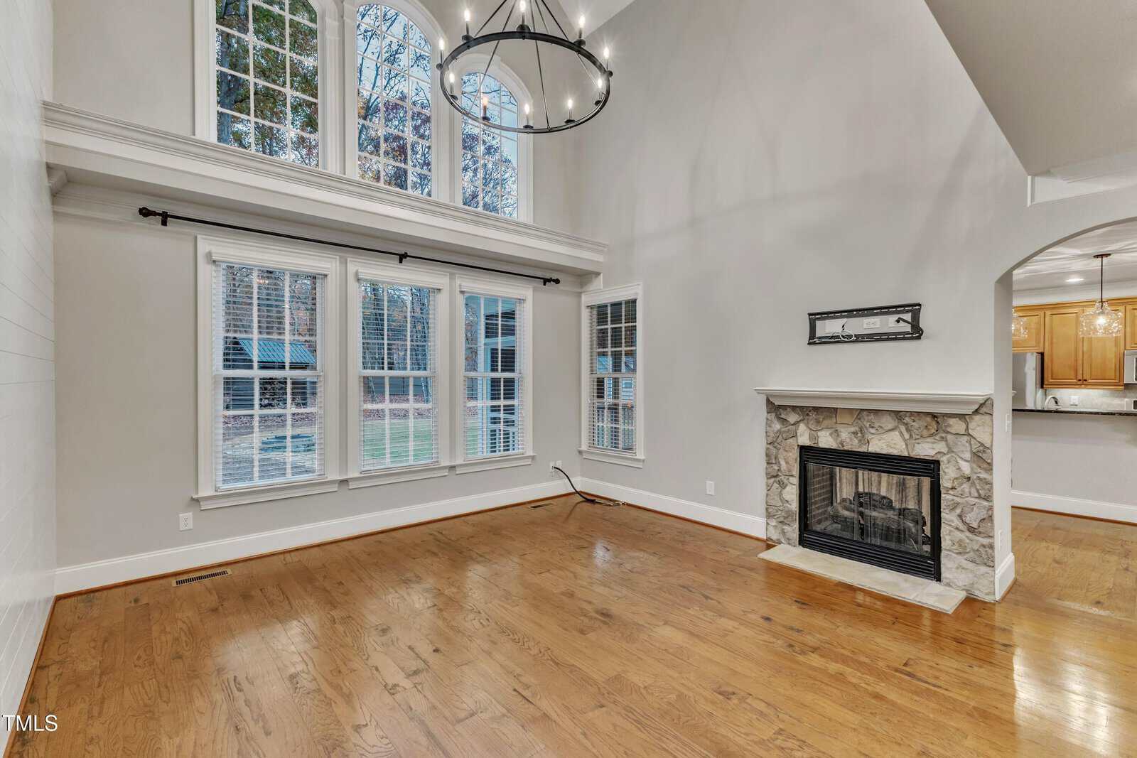 Spacious living room with large windows, high ceiling, wooden floor, stone fireplace, and a modern chandelier. The room receives ample natural light and has an open view into a kitchen area.