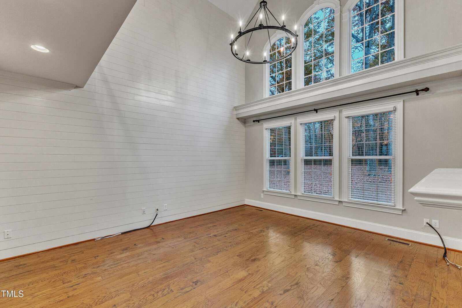 Spacious room with high shiplap walls, tall windows, hardwood floors, and a black chandelier. The room is empty, with white trim and natural light streaming in from the arched upper windows.