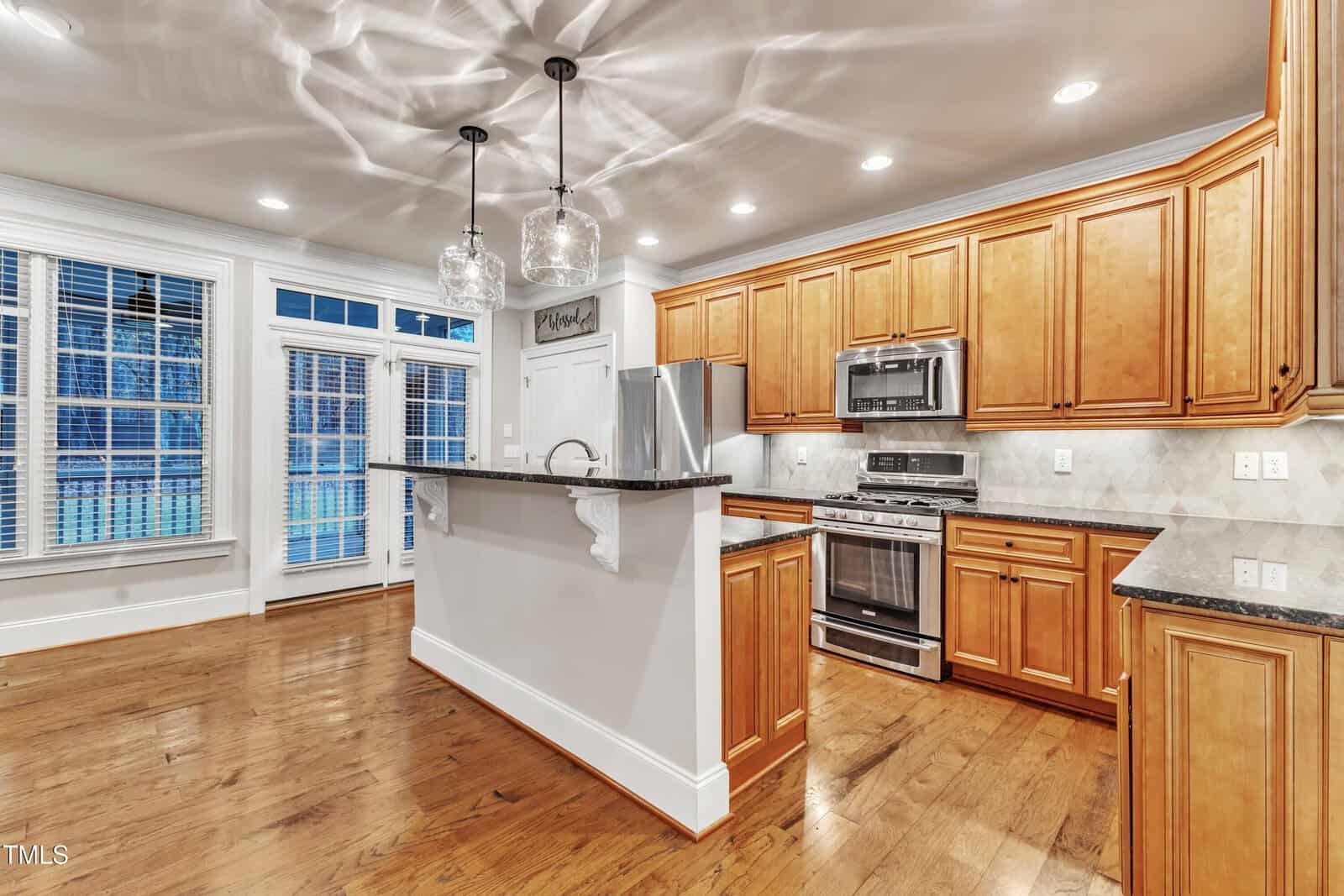 Modern kitchen with wooden cabinets, stainless steel appliances, granite countertops, pendant lights, hardwood floors, and large windows with white blinds. A bar-style counter separates the kitchen from the living area.