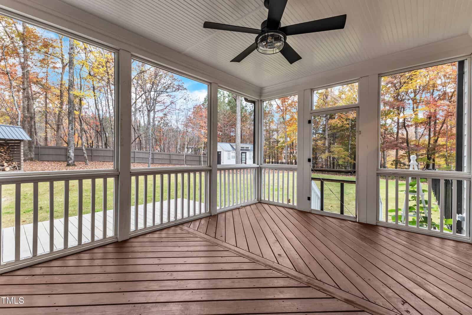 A screened-in porch with a wooden floor, white railing, and a black ceiling fan overlooks a grassy backyard with autumn trees and a small shed in the distance.