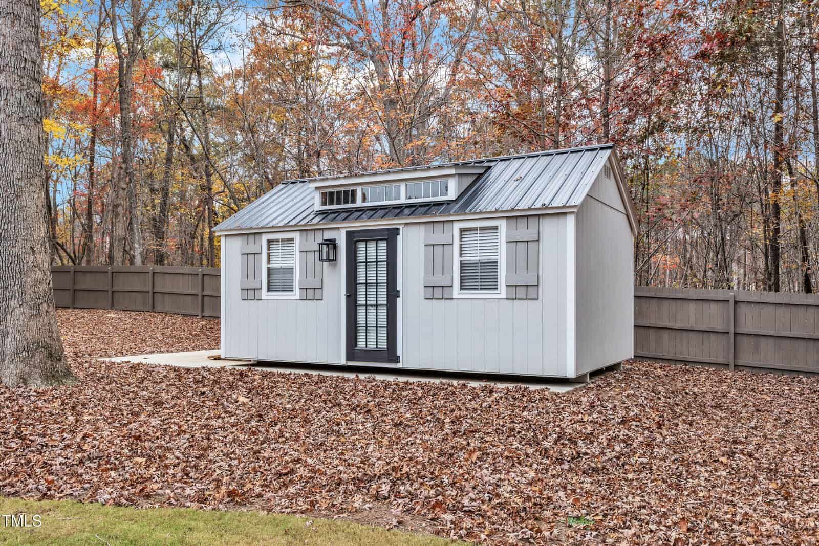 A small gray shed with white trim and a black door sits on a concrete pad in a fenced backyard covered in autumn leaves, with trees and a wooden fence in the background.