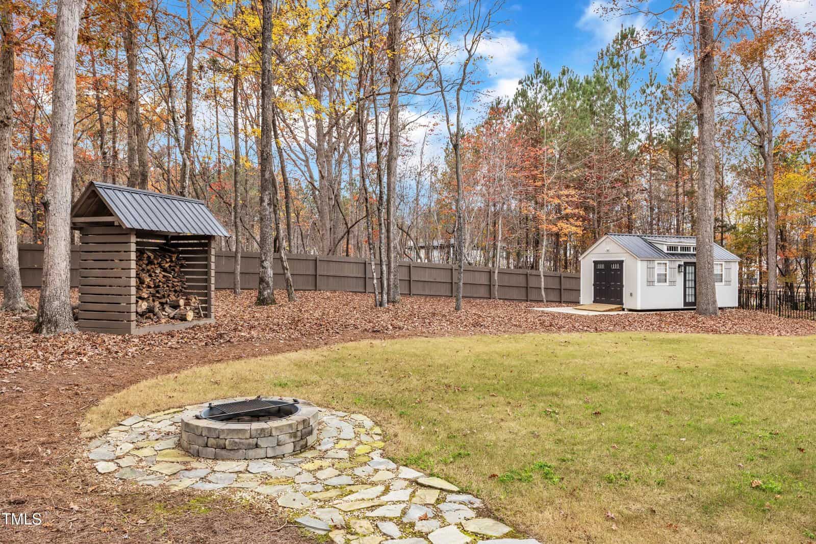 A backyard with a fire pit on a stone path, a wood storage shed, and a white shed. Trees with autumn leaves and a tall wooden fence enclose the yard under a blue sky with clouds.
