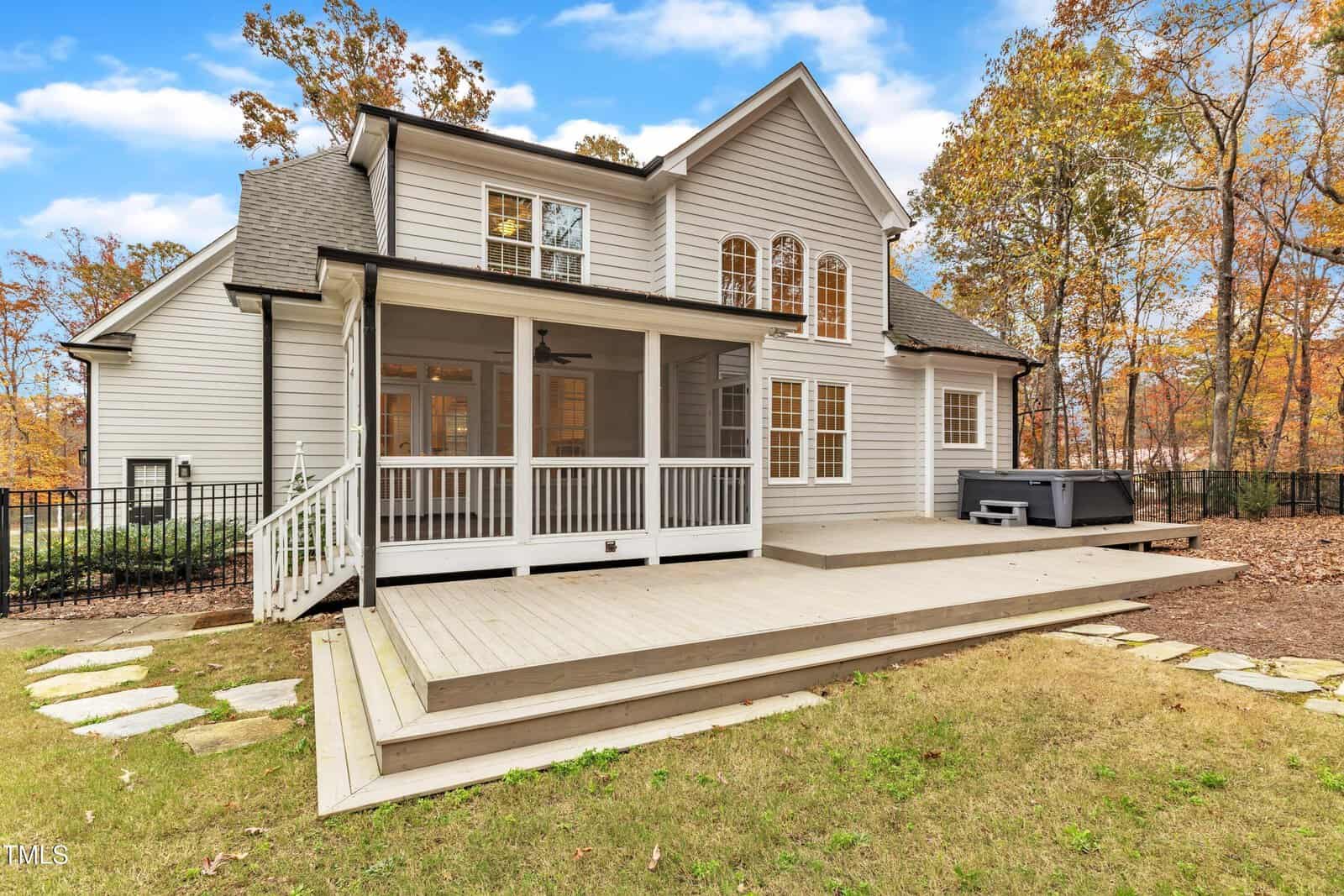 A two-story gray house with white trim features a spacious wooden deck, a screened-in porch, and a hot tub. The backyard has stone pavers and is surrounded by autumn trees.