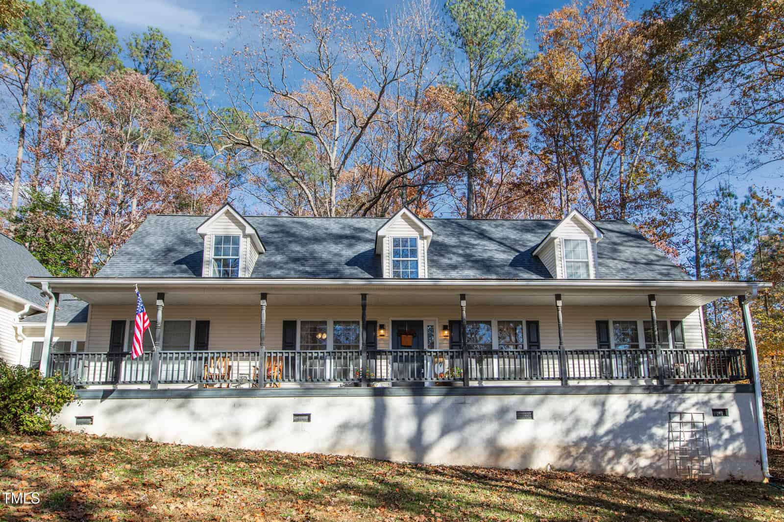 A two-story house with a large front porch, three dormer windows, white siding, and a gray roof, surrounded by autumn trees. An American flag hangs by the porch.