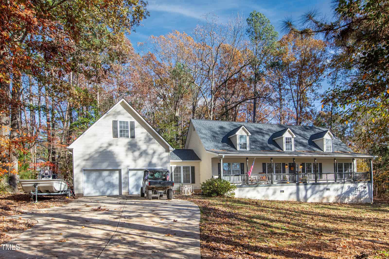 A two-story house with a large front porch and attached garage sits among tall autumn trees. A driveway leads to the garage, with a parked truck and a boat visible on the left side.