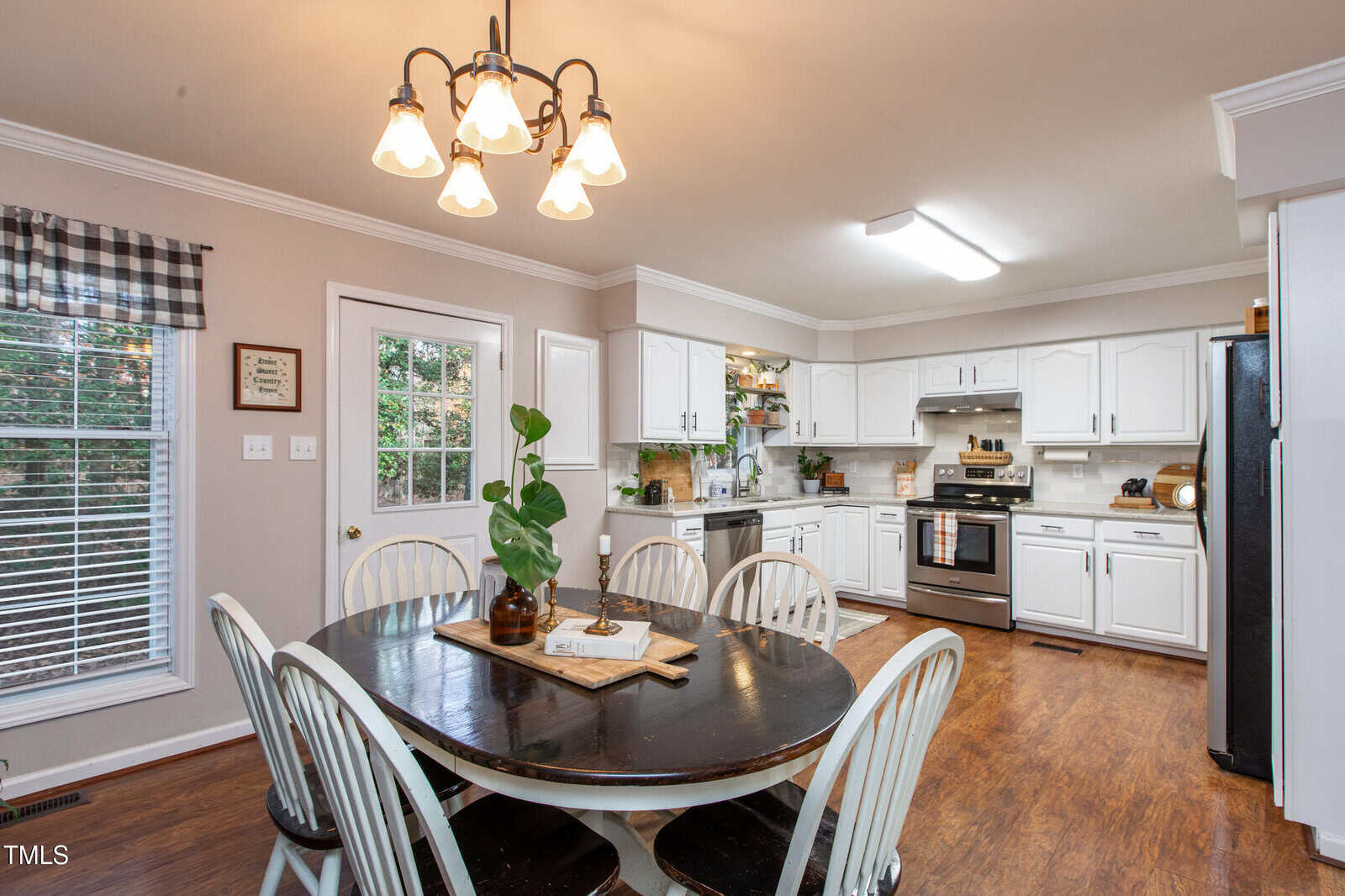 A cozy kitchen with white cabinets, stainless steel appliances, wood flooring, and a black dining table with white chairs. A window with checkered valance and a door lead to the outside. A plant decorates the table.
