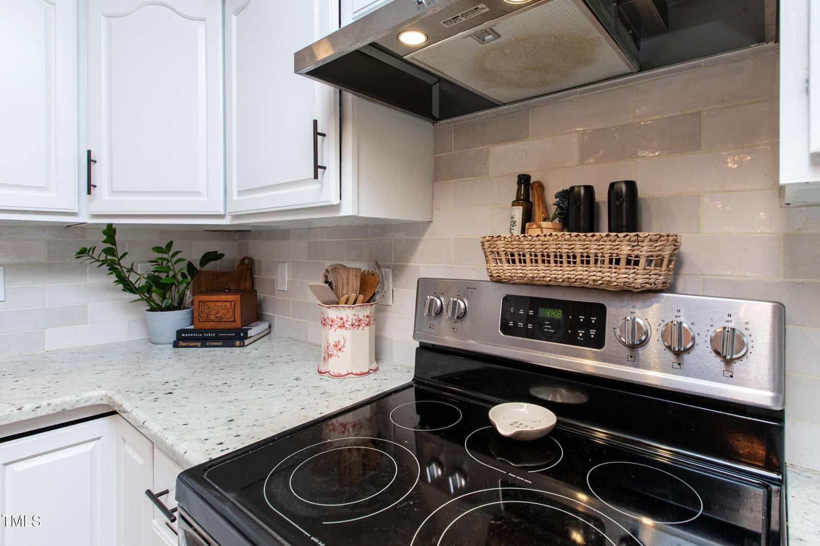 A modern kitchen corner with a stainless steel electric stove, white cabinets, marbled countertops, a potted plant, utensils in a ceramic jar, cookbooks, and a wicker basket holding bottles on a shelf above the stove.