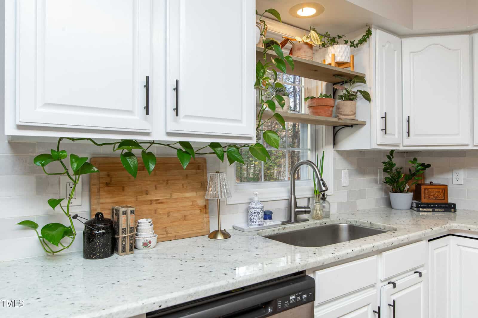 A bright kitchen with white cabinets and countertops, a plant vine draped along the counter and window, shelves holding potted plants and decor, and a window above the sink letting in natural light.