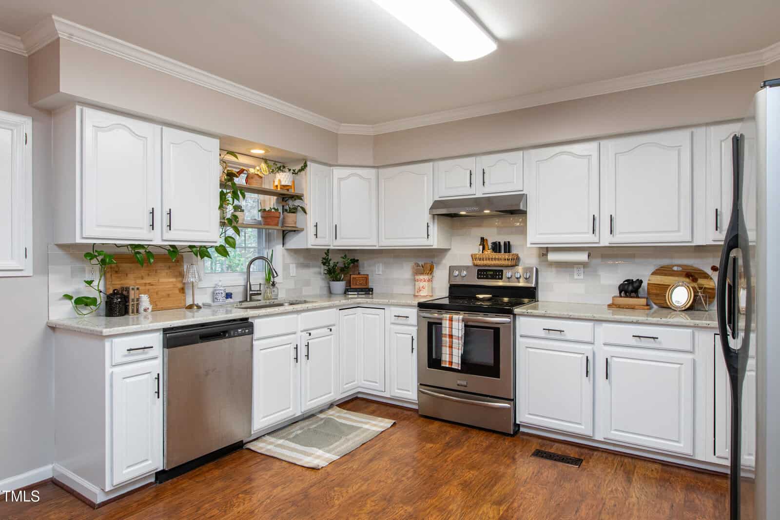 A modern kitchen with white cabinets, stainless steel appliances, wood flooring, and a small rug by the sink. Plants and kitchenware decorate the countertops, and under-cabinet lighting adds warmth.