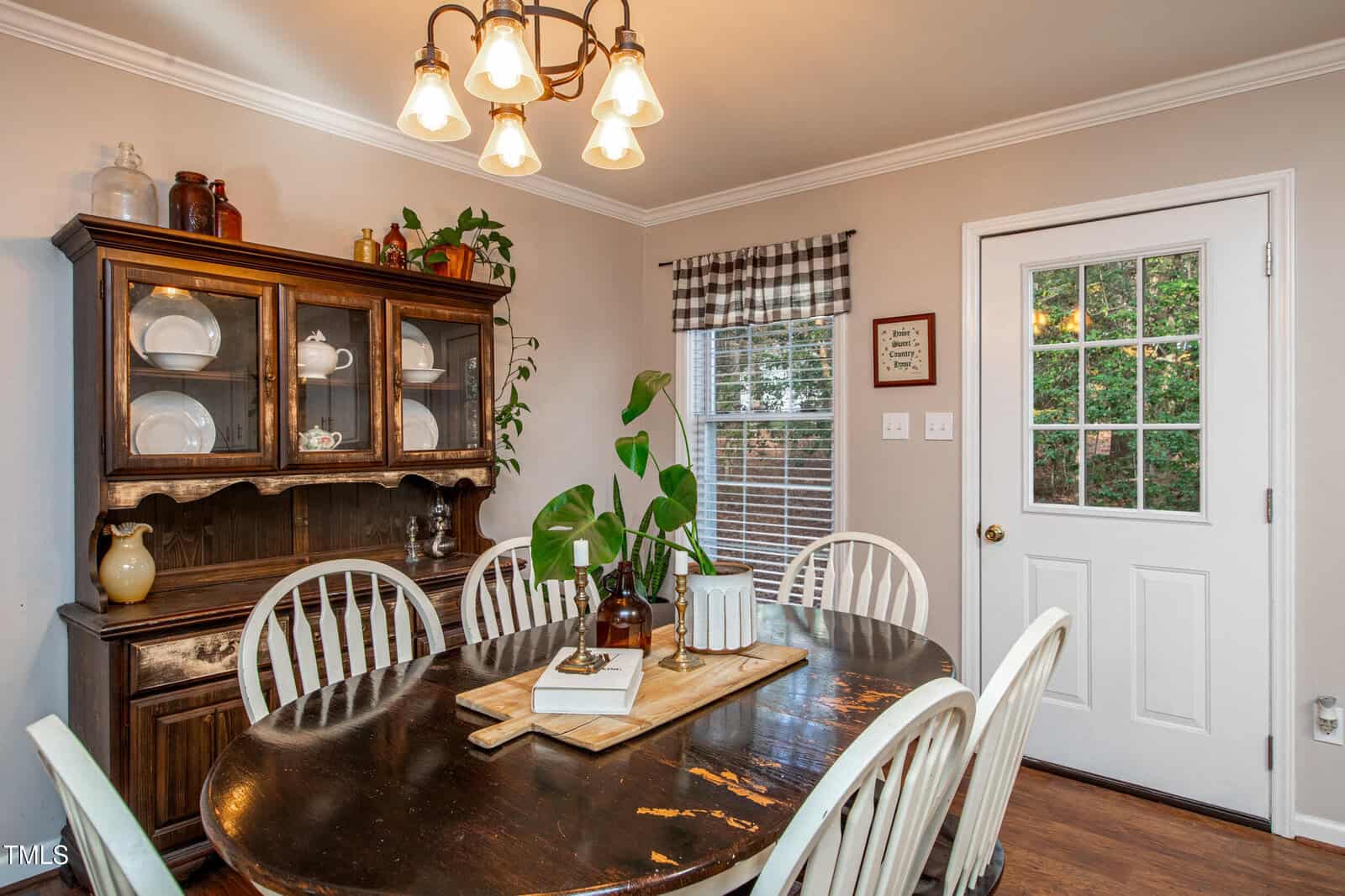 A cozy dining room with a wooden table, six white chairs, a potted plant, a hutch with dishes, a window with plaid curtains, and a door leading outside. Warm lighting hangs above the table.