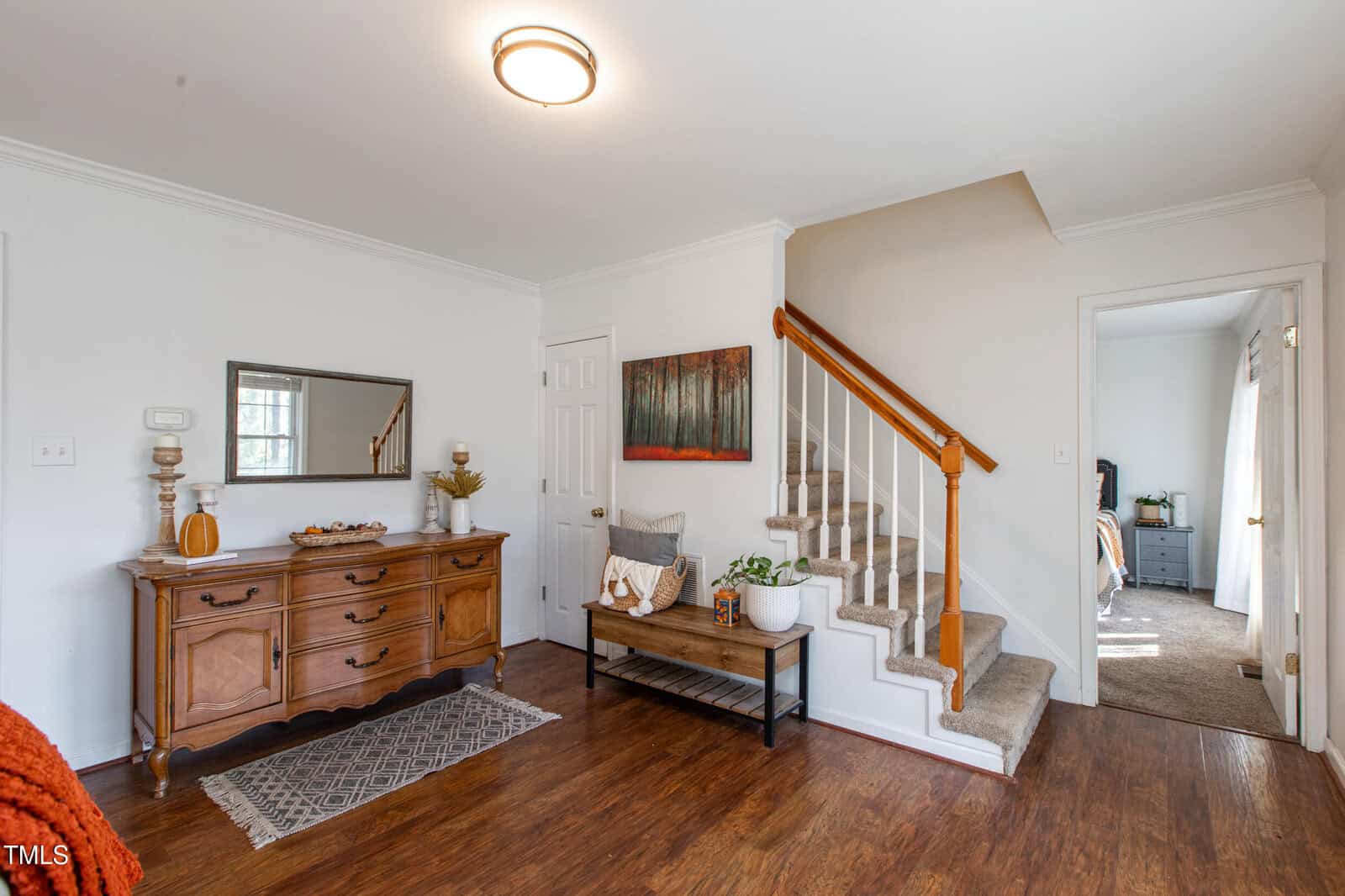A bright entryway with wood floors, a wooden sideboard with decor, a small bench with plants, a staircase with beige carpet, and a glimpse into a bedroom with sunlight.
