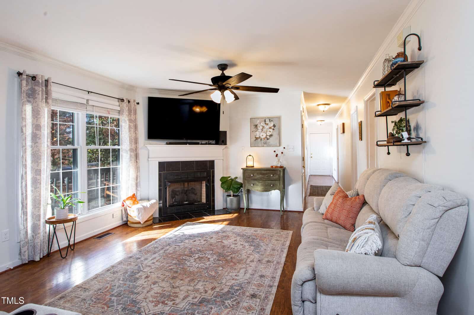 Bright living room with large windows, a ceiling fan, gray couch, patterned rug, fireplace with a TV above, plants, and shelves on the wall; wooden floors and a hallway leading to other rooms.