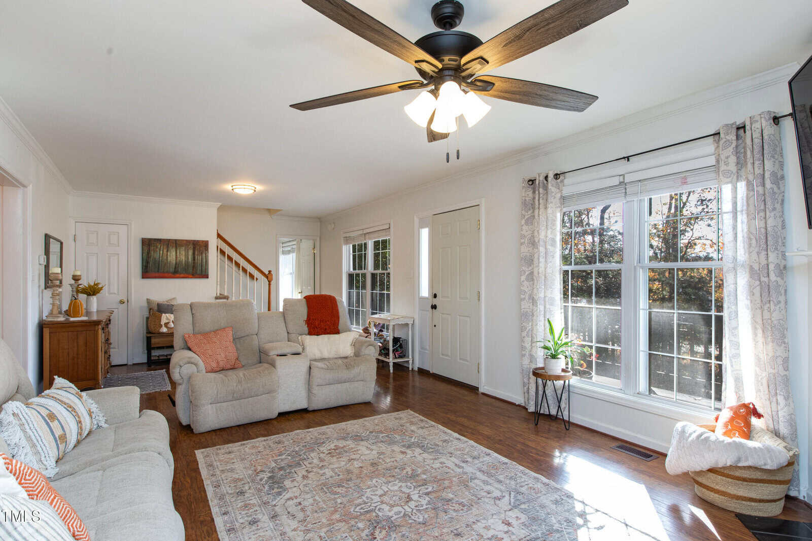 Bright living room with large windows, neutral walls, wood floor, a beige sofa and armchairs with orange accents, patterned rug, ceiling fan, houseplants, and sunlight streaming in through sheer curtains.