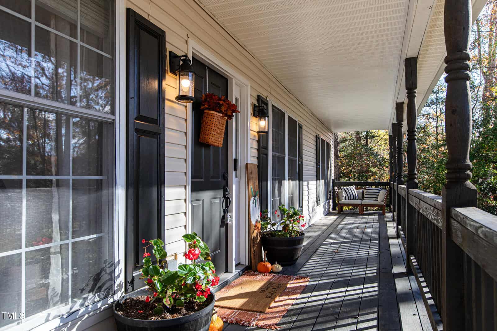 A sunlit front porch with black railings and shutters, potted plants, a hanging basket, pumpkins, and a welcome mat. A wooden swing sits at the far end, and autumn trees are visible in the background.