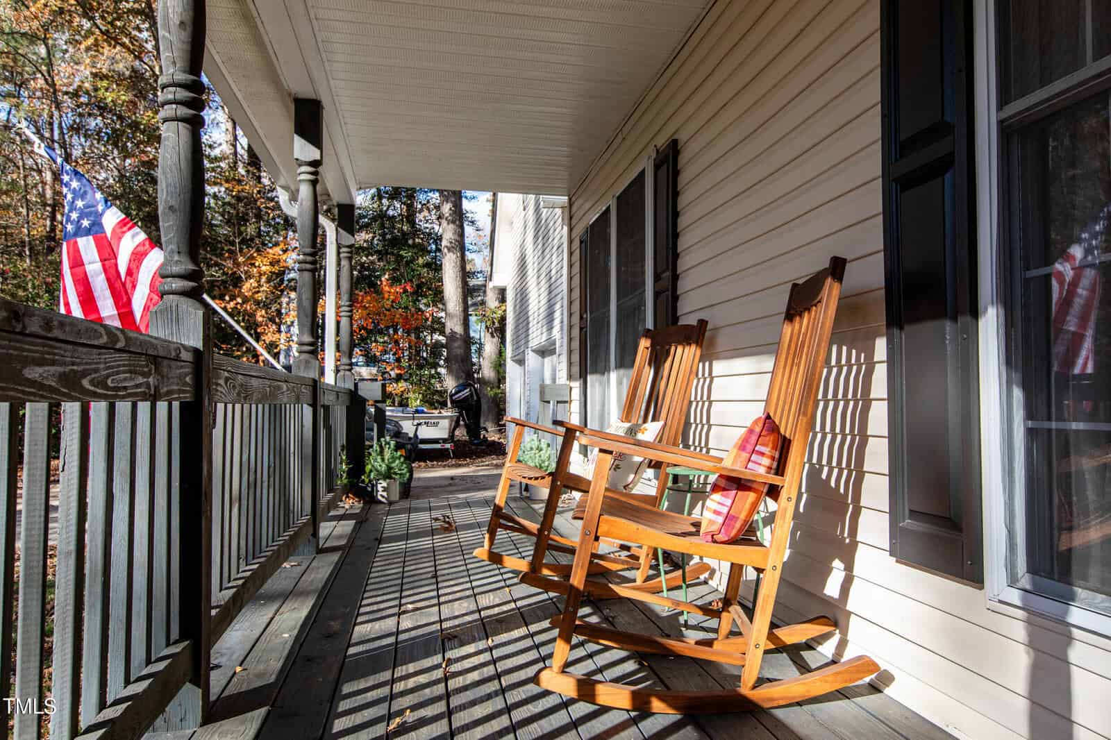 A sunlit porch with two wooden rocking chairs, a pillow, and an American flag hanging from the railing, overlooking a yard with autumn trees. Shadows from the railing stretch across the floor.
