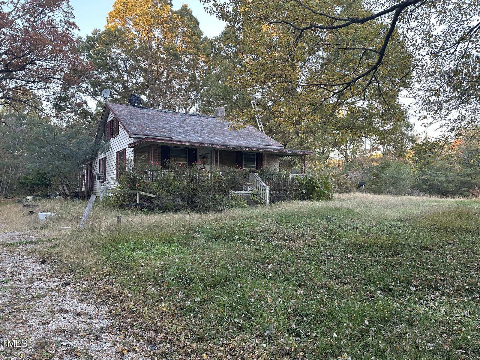 A weathered, single-story house with a slanted roof, surrounded by overgrown grass and trees. It has a porch with peeling paint and some foliage growing around it. The background includes tall trees with autumn leaves. A gravel path leads to the house.