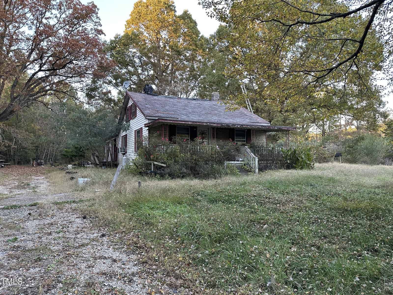 A small, weathered house with a porch is surrounded by overgrown grass and trees with autumn leaves. A gravel path leads to the house, which appears to be in a rural area during the fall season.