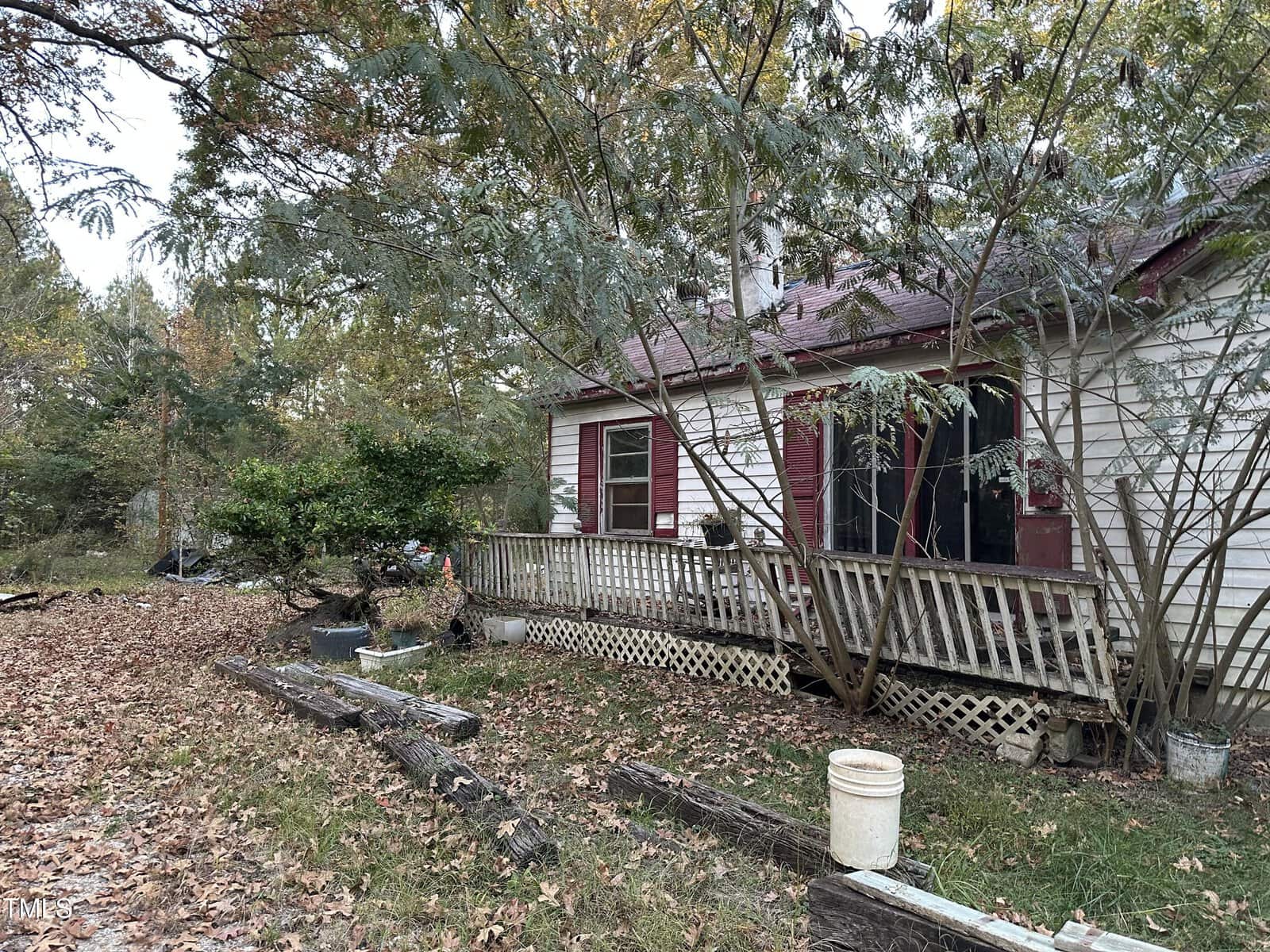 A small, worn-out house with a porch is surrounded by trees and overgrown foliage. The wooden railing and steps show visible wear. Fallen leaves cover the ground, and a few items, including a white bucket, are scattered near the porch.
