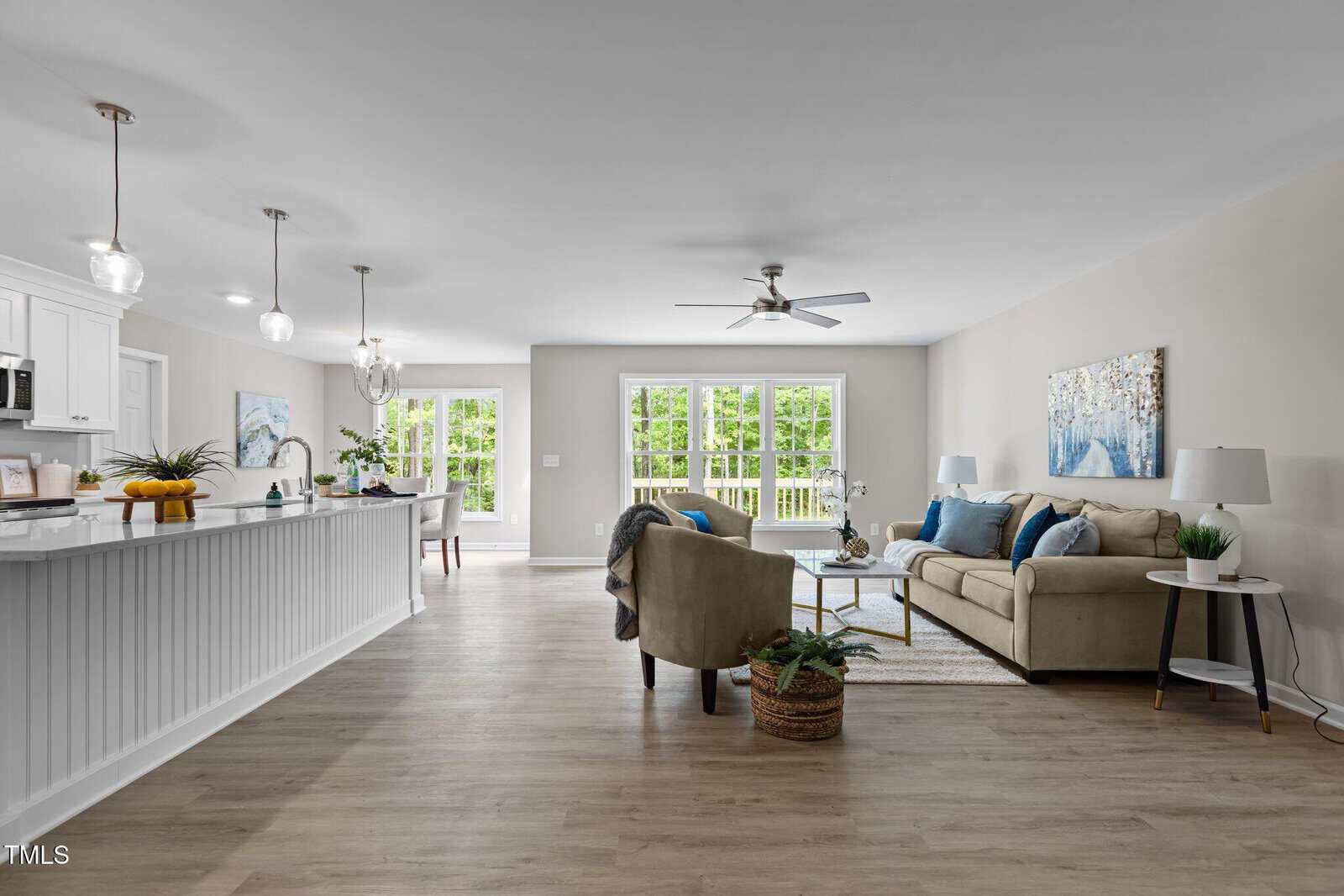A modern open-concept living space with a kitchen island, white cabinetry, light wood floors, a beige sofa, armchair, coffee table, and large windows overlooking a green backyard.