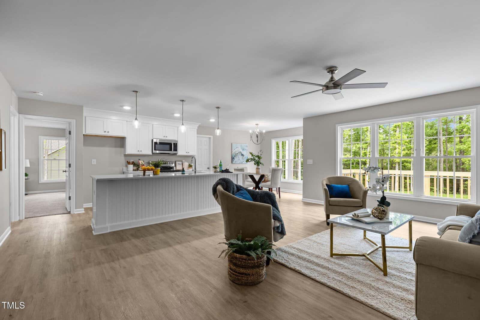 Open-concept living area with wood flooring, a modern kitchen with white cabinets and stainless steel appliances, a dining space, and large windows providing natural light. Two armchairs and a couch face a glass coffee table.